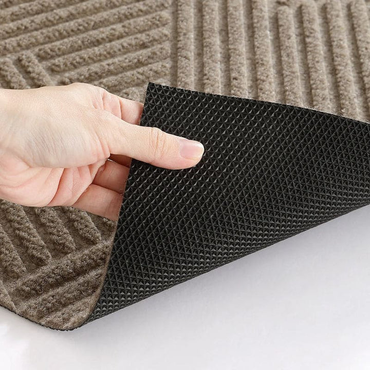 Hand lifting corner of beige doormat to show non-slip black rubber backing texture.