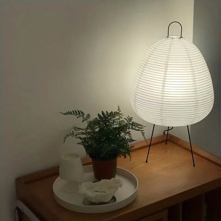 Warm white AKARIS rice paper lantern table lamp with black tripod legs on a wooden sideboard, ideal for bedroom lighting