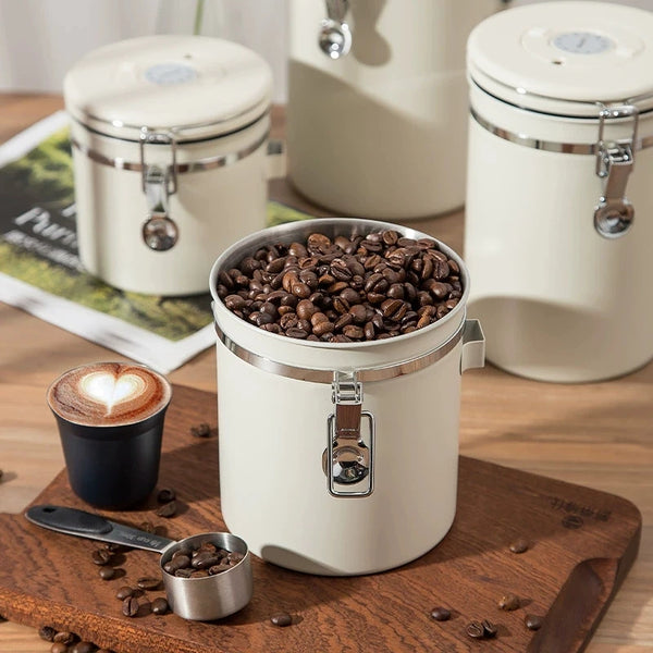 Cream airtight coffee canister filled with beans beside a cup of coffee on wooden surface.