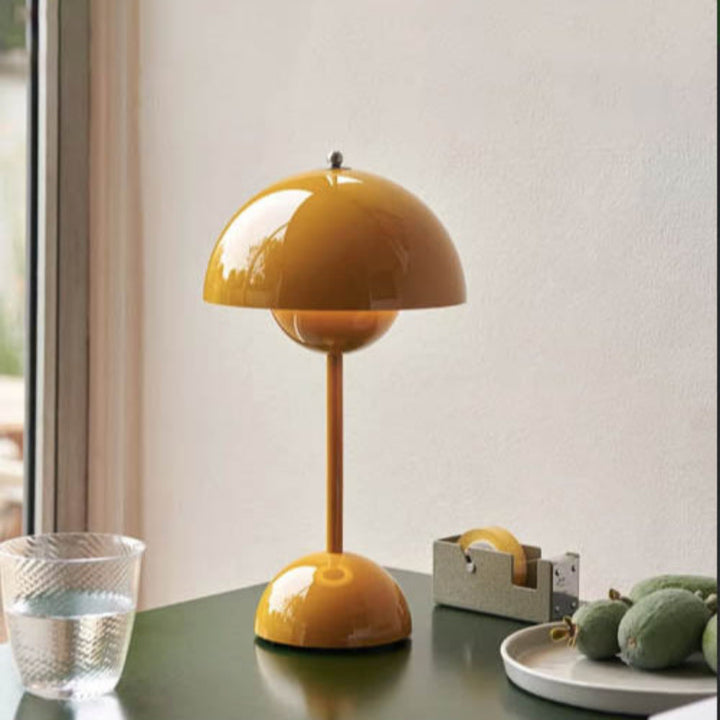 Yellow rechargeable dome table lamp placed on a green side table with a glass of water, fruits, and minimalist office accessories in a sunlit room.