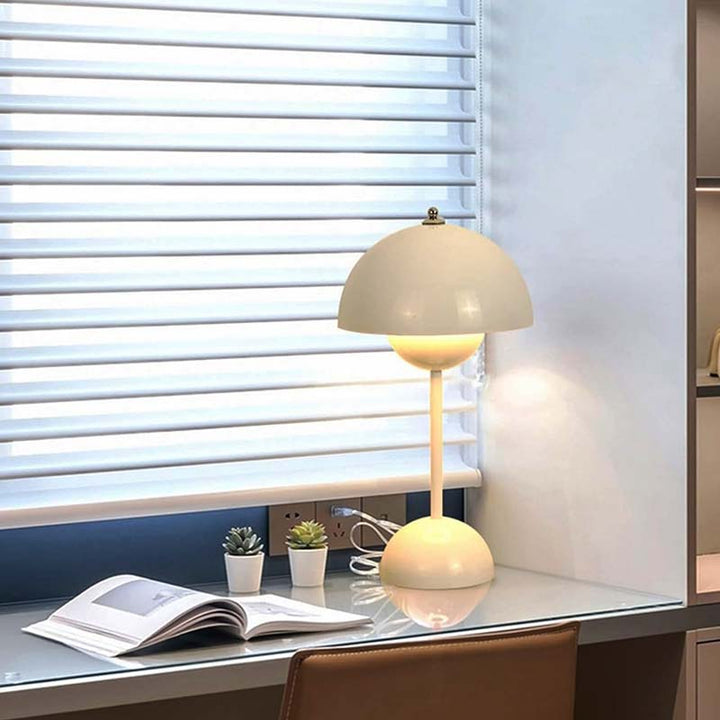 Modern flower bud table lamp with dome shade and spherical base, placed on a glass desk near window blinds for stylish home office lighting.