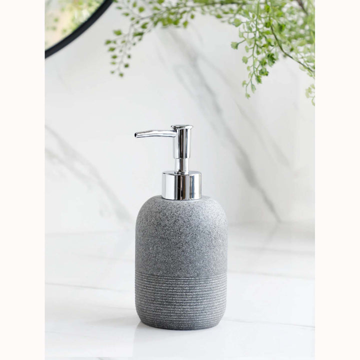 Gray textured soap dispenser with silver pump on a white surface with a blurred green plant in the background