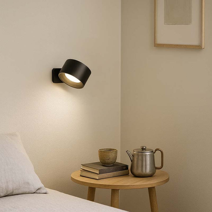 Black wall lamp on a beige wall with a wooden side table holding a teapot and books.