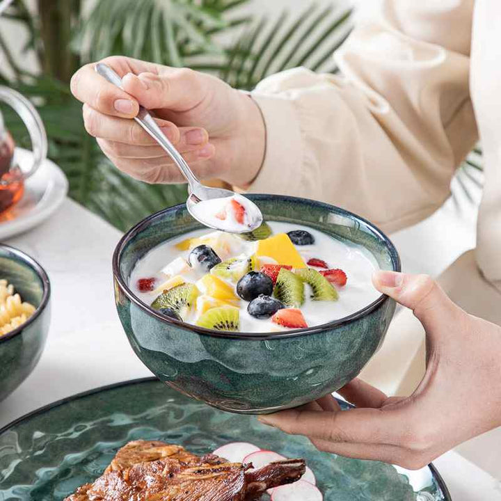Person holding a bowl of fruit salad with yogurt, surrounded by greenery.