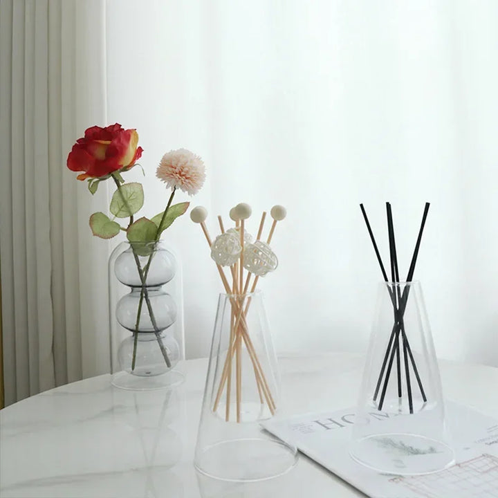 Two transparent glass essential oil diffusers with reed sticks beside fresh flowers on white table