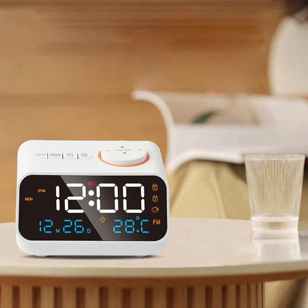 White digital FM Radio LED Alarm Clock placed on a bedside table beside a glass, showing time, date, and temperature.