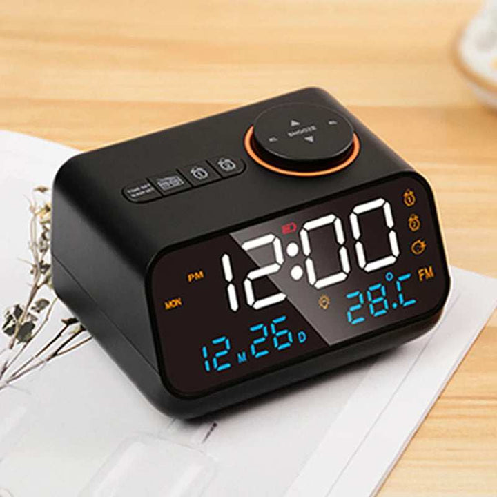 Black FM Radio LED Alarm Clock with large LED display and orange snooze button placed on desk with papers.