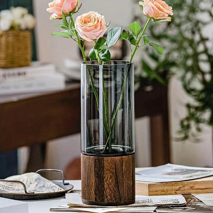 A tall, slim, colored glass propagation vase with a wooden base, displayed on a table with pink flowers inside it.