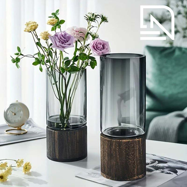 Two cylindrical vases with wooden bases on a table, one containing flowers.