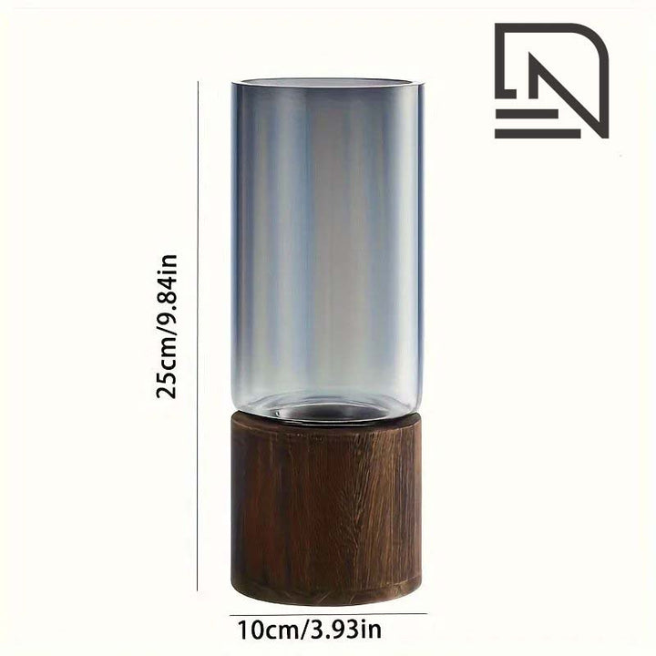 A tall, slim, colored glass propagation vase with a wooden base, displayed on a table with pink flowers inside it.