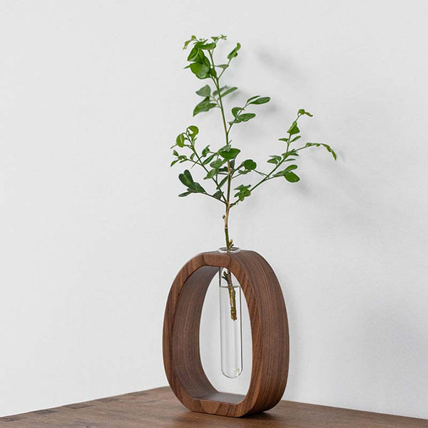 Wooden flower vase with a circular design holding a clear glass tube with a plant on a wooden surface.