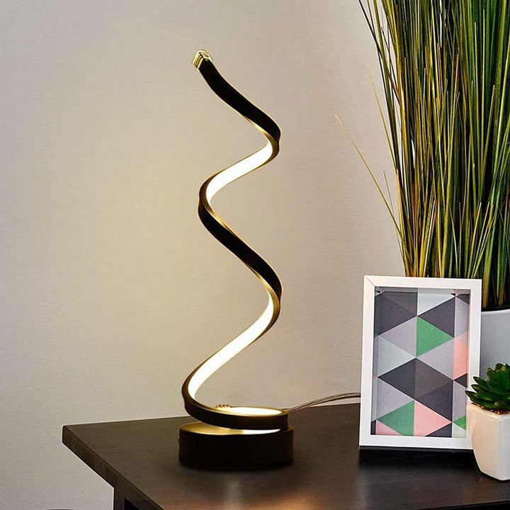 Modern night light with a spiral design on a desk next to a plant and framed picture.