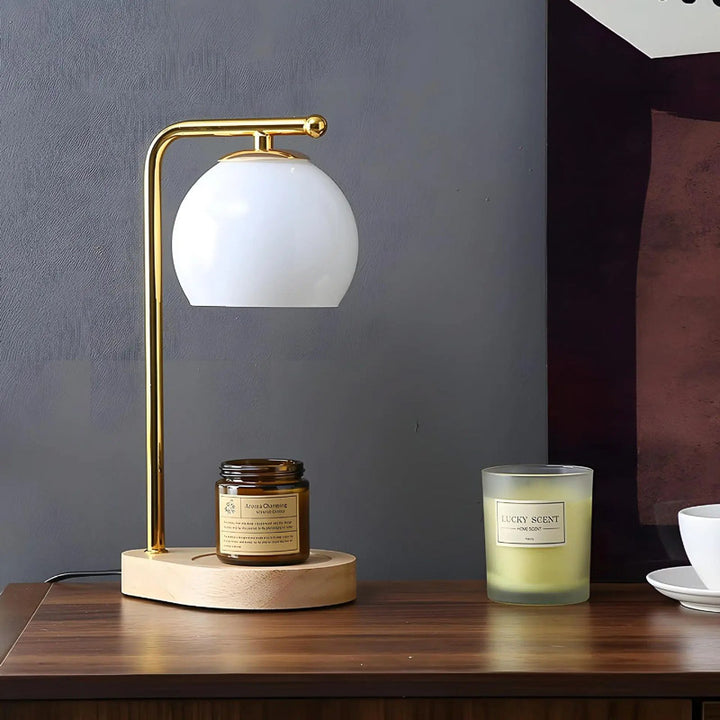 Table lamp with a white glass shade on a wooden base, accompanied by candle warmer and a a cup on a wooden surface.