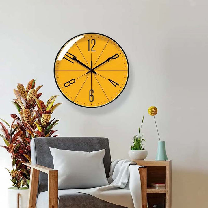 Yellow wall clock on a white wall above a gray armchair with a plant and small table in the foreground.