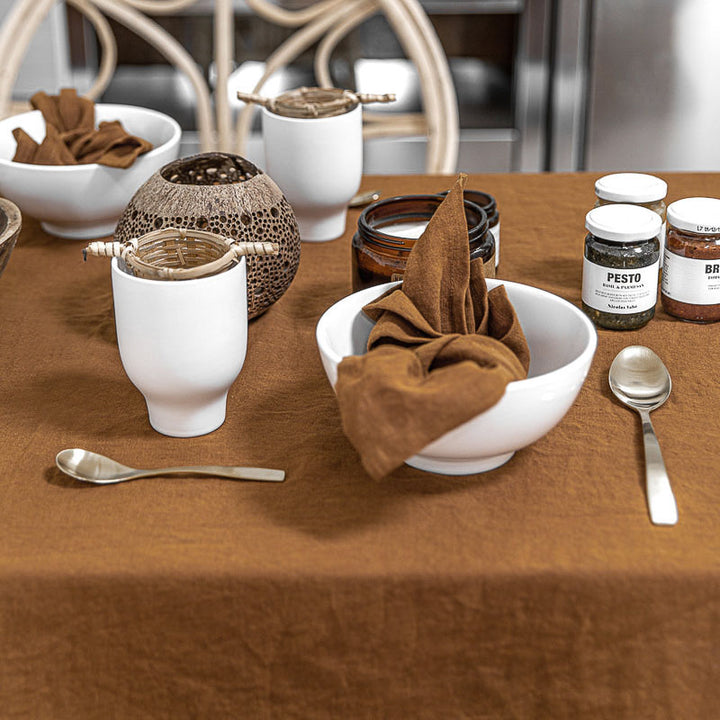 Table setting with ceramic bowls, jars, and utensils on a brown - cinnamon linen tablecloth.