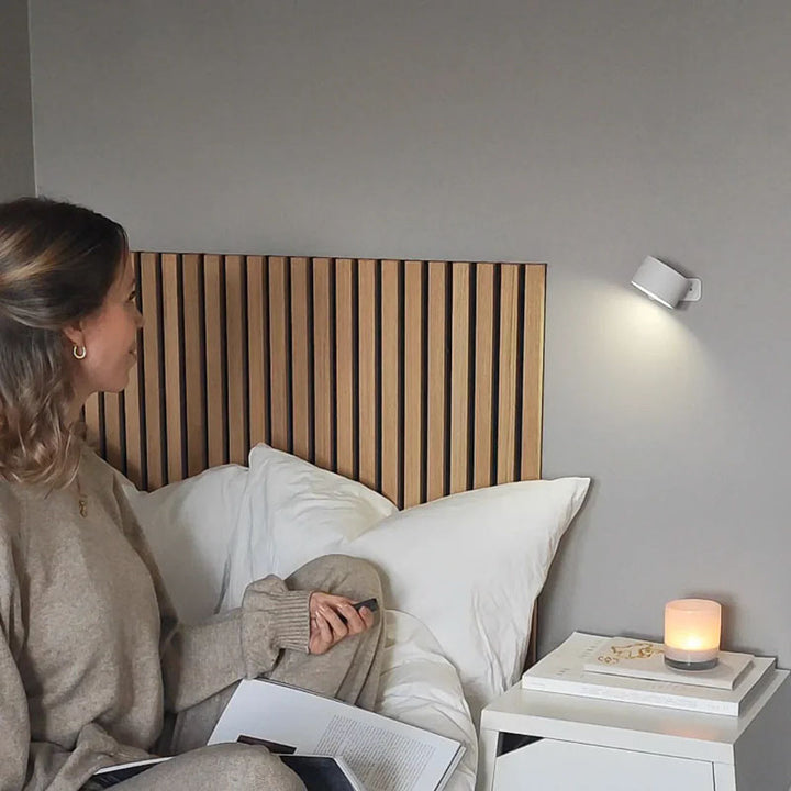 Woman sitting on a bed with a wooden headboard, reading a book, in a room with a nightstand and candle.