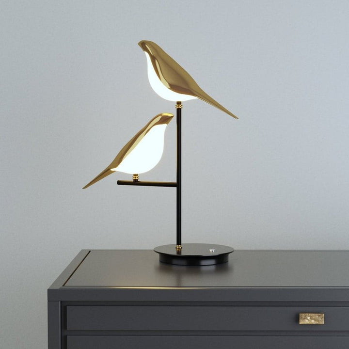 Modern golden bird LED table lamp on black stand, illuminating contemporary interior.