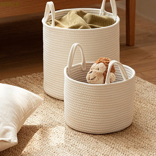 Two beige solid woven laundry baskets with handles, one empty and one containing a stuffed toy, placed on a floor with a pillow beside them.