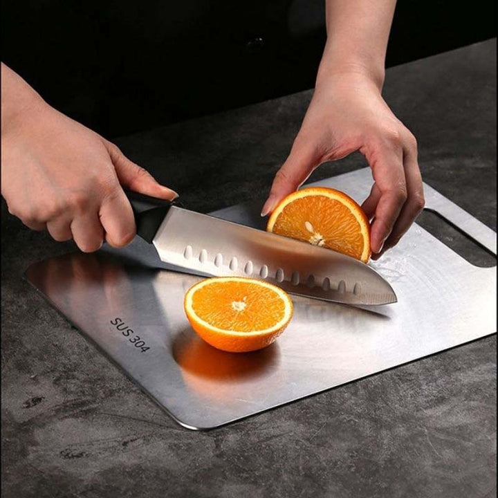 Person cutting an orange with a knife on a stainless steel cutting board.