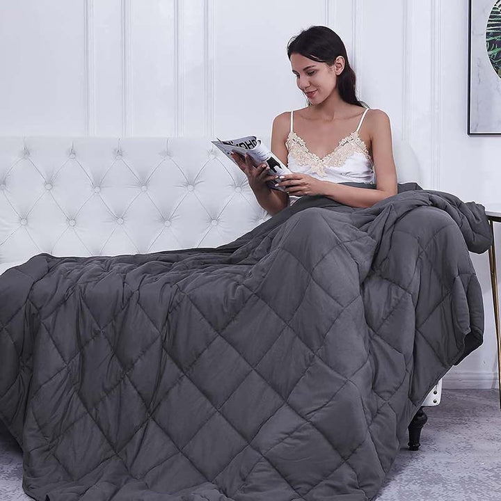 Woman reading a book under a grey weighted blanket quilt in a bedroom setting