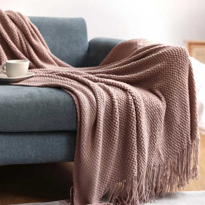 Muted pink acrylic waffle-knit throw with tassel fringe spread across a sofa for soft neutral interior accents