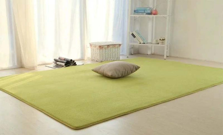 Green rug with a pillow on a light wooden floor in a room with white curtains and shelves.