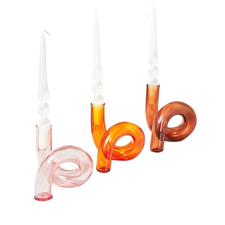 Pink, orange, and amber curved glass candle holders each styled with white spiral candles on clean white surface