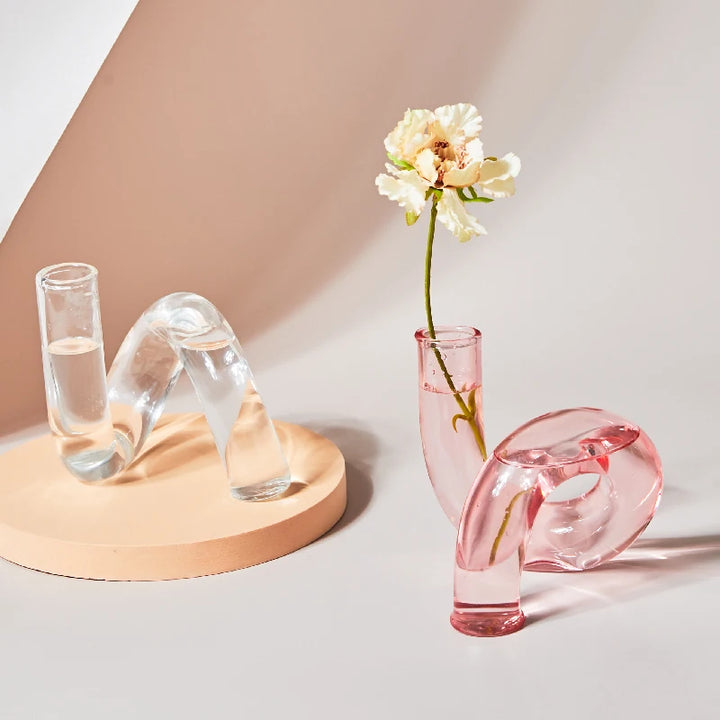 Clear and pink glass flower vases with curved design holding water and single white blossom on pastel surface