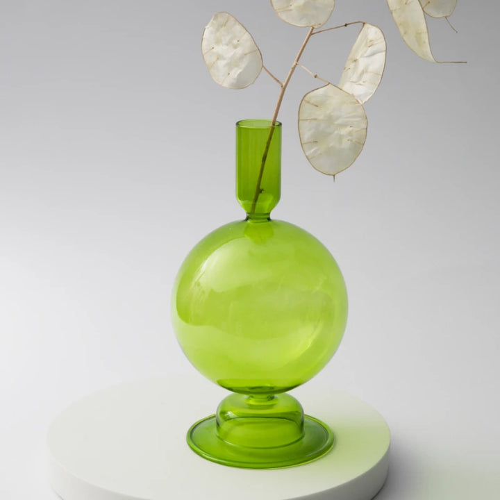 Green glass round candle holder with narrow neck used as vase holding dried branches on white stand