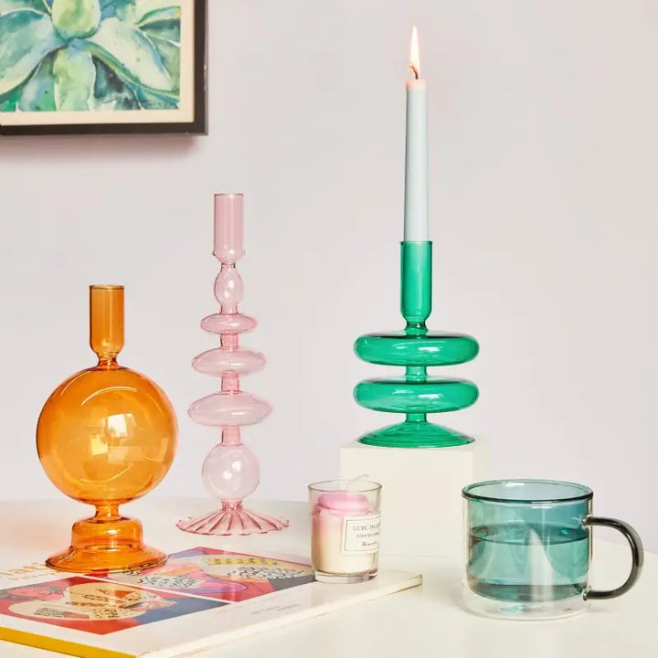 Colorful glass taper candle holders in amber, pink, and green displayed on white table with books and glassware