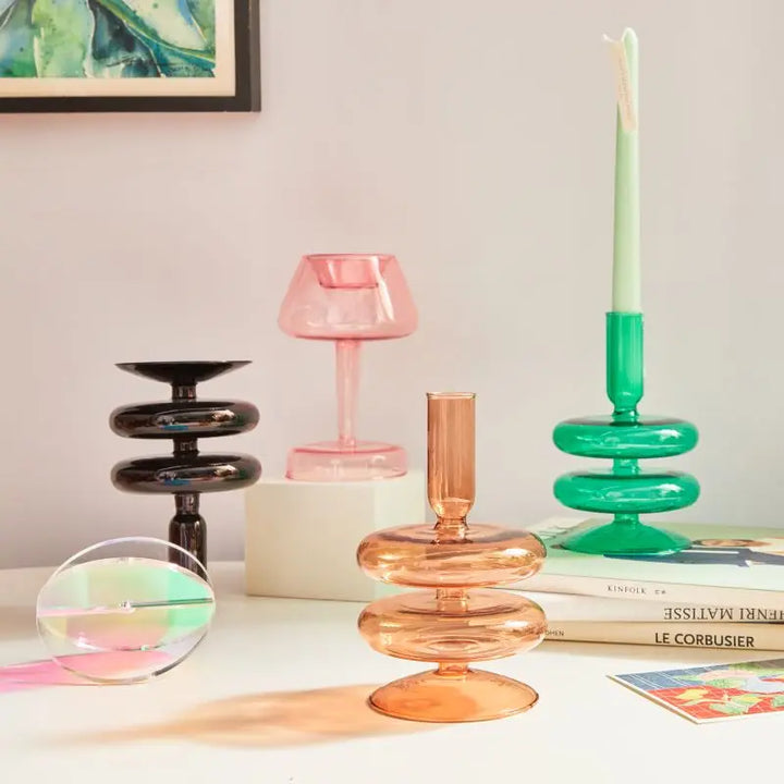 Modern glass candle holders in green, amber, pink, and black arranged on table with pastel decor in Nordic style