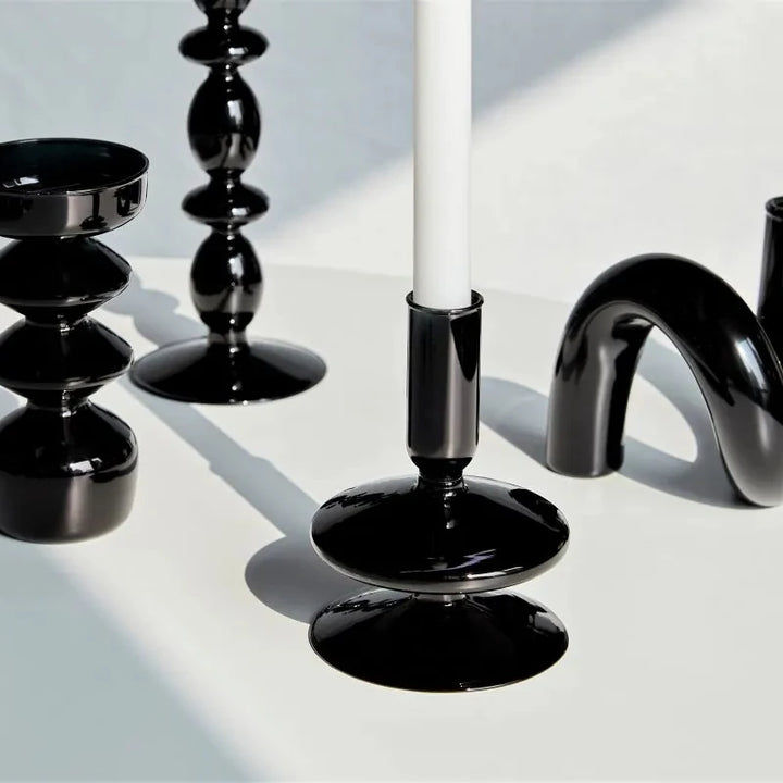 Glossy black glass candle holders in various sculptural shapes with white taper candle on minimalist surface