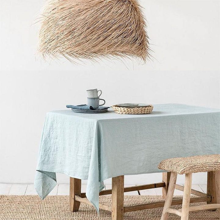 Table with a light blue linen tablecloth, cup, and saucer, with a woven chair and pendant light in a minimalistic room.