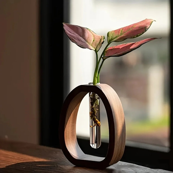 A minimalist walnut and glass mini flower vase placed on a table, with a clear glass tube in the center containing a hydroponic plant.