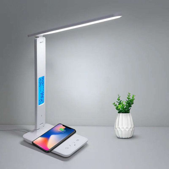 White LED desk lamp charging a smartphone wirelessly beside a small ceramic vase on a desk