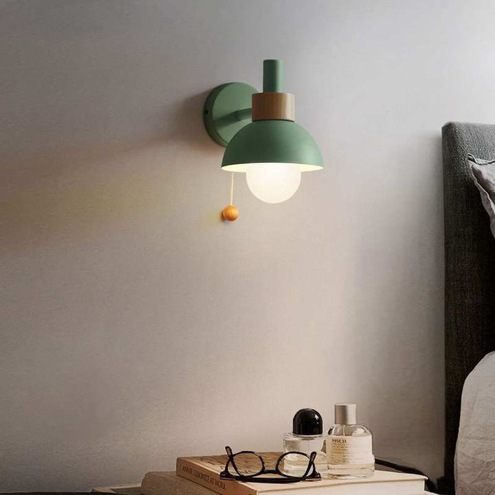 Green wall lamp with wood accent casting warm light beside a bed in Nordic modern style.
