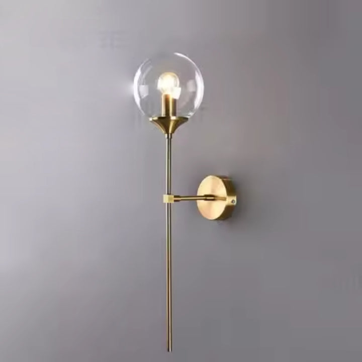 Clear glass globe wall lamp with brushed brass base, offering soft ambient lighting against a grey wall.
